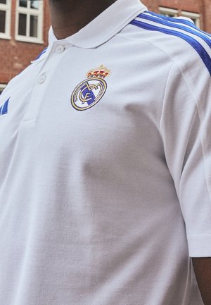 Person trägt weißes Poloshirt mit blauen Streifen und Real Madrid-Logo auf der Brust, steht draußen in der Nähe eines Ziegelgebäudes.