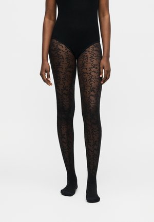 Woman wearing sheer black patterned tights and a black bodysuit, standing against a plain light background.