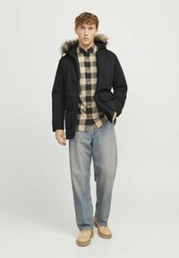 Sort pufferjakke med faux fur-forsynet hætte, gråternet skjorte og lyseblå jeans. Beige sko fuldender outfittet.