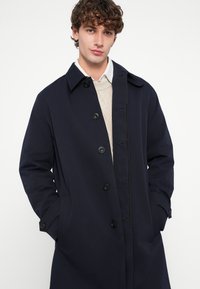 Tmavomodrý trenčkot vyrobený z hladkej látky, s gombíkovým zapínaním vpredu, veľkým golierom a bočnými vreckami. Jemné švy ako akcenty.