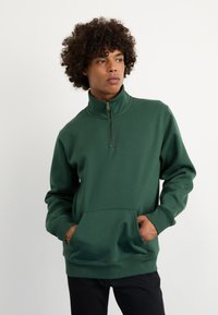 Zeleni zip-up dukserica napravljena od mekog materijala. Ima visoki ovratnik, prednji džep i rebraste manžete. Jednostavan, jednobojan dizajn.