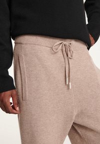 Beige Strick-Jogginghosen verfügen über einen elastischen Bund mit Kordelzug, Seitentaschen und strukturierter Stoff. Tragen Sie dazu einen schwarzen Pullover.