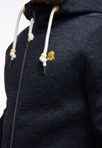 Granatowy, rozpinany na suwak polar z kapturem, wykonany z teksturowanej tkaniny, z białymi sznurkami do ściągania ozdobionymi drewnianymi koralikami oraz małym żółtym logo na piersi.