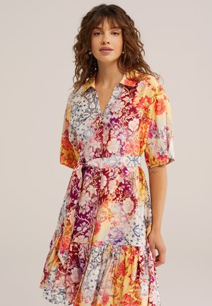 Hölgy hullámos barna hajjal, övvel megkötött, többszínű virágos ruhát visel rövid ujjú, egyszínű háttér előtt.