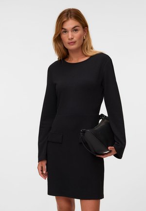 Femme aux cheveux châtain clair portant une robe noire à manches longues, tenant un sac à main noir devant un fond clair uni.