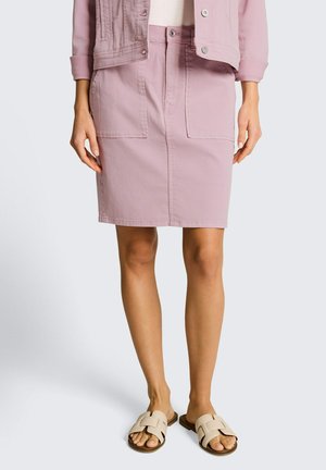 Vrouw draagt een lichtroze denim rok met voorzakken, bijpassend jasje, witte top en beige slippers, staand tegen een effen achtergrond.