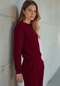 Felpa bordeaux con scollatura rotonda e polsini a costine, abbinata a pantaloni jogger coordinati. Il tessuto sembra morbido con una texture liscia.