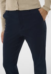 Tumed sinine kohandatud püksid sileda tekstuuriga, millel on eesmistest taskud ja kitsas lõige, mida täiendab beež kardigan.