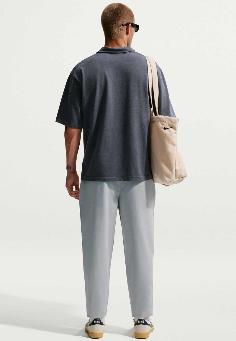 Uomo con polo grigio scuro, pantaloni grigio chiaro, occhiali da sole neri, sneaker Nike bianche, porta una borsa Nike beige sulla spalla, visto di spalle.