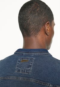 Denim jakna v temno modri barvi, klasičnega kroja, z logotipom na zadnjem delu. Utrjena tekstura z kontrastnimi šivi.
