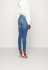 Högmidjade blå denimjeans med smal passform, med bakfickor, två fickor och en logotyp på midjebandet, i kombination med vita klacksandaler.