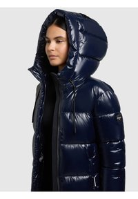 Tumši zila pūtuma jaka ar spīdīgu apdari, lielizmēra kapuci, priekšējo rāvējslēdzēju un lielām šūtām daļām. Piedāvā piedurknes logo plāksteri.