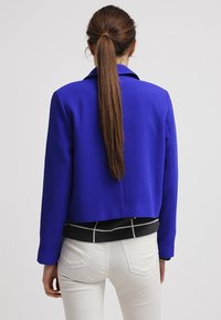 Blazer blu tagliato realizzato in tessuto texture, con colletto e maniche lunghe, indossato sopra una camicia a righe bianco e nera e pantaloni bianchi.