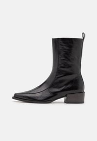 Bota de tobillo de piel negra con punta afilada, silueta estructurada y un bajo tacón bloque. Presenta una superficie lisa y una tira lateral para halar.