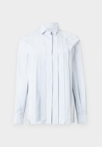 Svetlo modra dolga majica s ovratnikom v obliki konice, z navpičnimi naborki spredaj in zapenjanjem s gumbi pri vratih. Gladka tekstura tkanine.