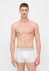 Boxer-briefs blancs avec une ceinture élastique grise portant le logo "GUESS". Le tissu semble lisse, ajusté et extensible.