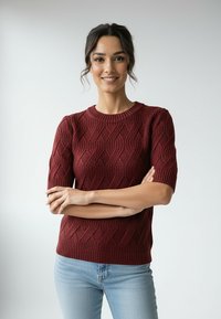 Bordowy sweter dzianinowy z krótkimi rękawami, z teksturowanym falistym wzorem. Do kompletu jasnoniebieskie dżinsy.