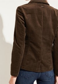 Ruskea puuvillakorduroy-takki, jossa istuva leikkaus, lovellinen kaulus ja hillitty tekstuuri. Takissa on keskisauma ja pitkät hihat. Yhdistetty sinisiin farkkuihin.