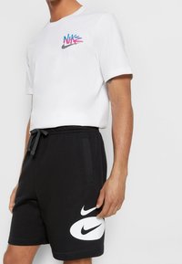 T-shirt en coton blanc arborant un logo Nike coloré, associé à un short noir affichant un grand swoosh Nike blanc. Vêtements de sport décontractés.
