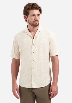 Man met kort bruin haar en baard draagt een beige, met patroon bedrukte, kort mouwig overhemd met knopen en olijfgroene broek, kijkt iets naar rechts tegen een witte achtergrond.
