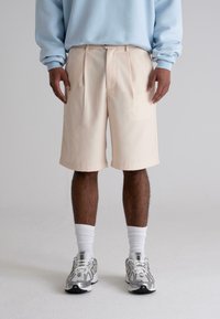 Beige pleated shorts with a tailored design, made from lightweight fabric, paired with white socks and grey sneakers.