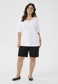 T-shirt blanc à col en V avec des manches courtes à motifs, associé à un short noir et des chaussures plates. Tissu doux, coupe décontractée et couleurs neutres.
