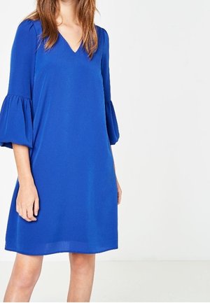 Kobieta ubrana w królewsko niebieską sukienkę do kolan z dekoltem w kształcie litery V i rękawami o długości do łokcia zakończonymi dzwonkami na jednolitym białym tle.