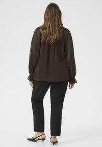 Mujer con pelo largo castaño, vistiendo una blusa sheer marrón oscuro, pantalones negros ajustados y tacones kitten de tira negra, de pie de espaldas.