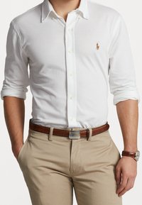 Vit långärmad skjorta med knappar av bomull med krage, prydd med en liten polo-spelare logo, i kombination med tanbyxor och ett brunt bälte.