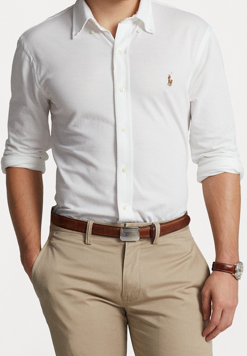 Vit långärmad skjorta med knappar av bomull med krage, prydd med en liten polo-spelare logo, i kombination med tanbyxor och ett brunt bälte.