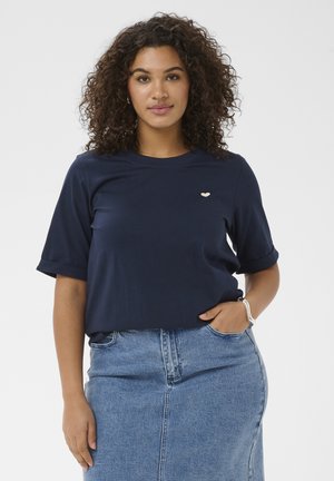 Femme aux cheveux bouclés portant une chemise à manches courtes bleu marine et une jupe en jean taille haute, main dans la poche, sur fond blanc.