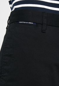 Pantaloni in cotone neri con una texture liscia, una vestibilità slim e un'etichetta in vita con la scritta "Scotch & Soda". Maglietta a righe parzialmente visibile.
