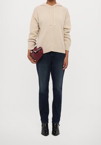 Personne portant un pull à capuche beige, un jean bleu foncé, des bottines noires, tenant un petit sac à main bordeaux sur un fond blanc uni.