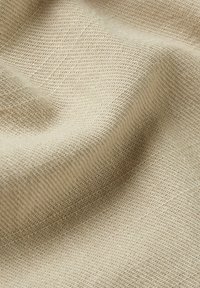 Beige stof met een gestructureerd, geweven patroon. Het oppervlak lijkt zacht en iets rekbaar, waardoor subtiele plooien en golven ontstaan.