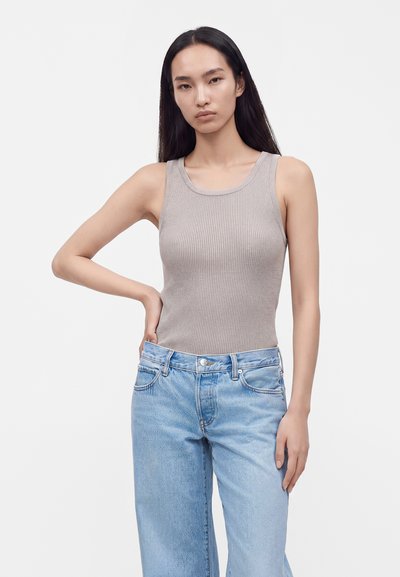 Jeune femme aux longs cheveux foncés portant un haut gris sans manches en maille côtelée et un jean bleu clair, debout avec une main sur la hanche.