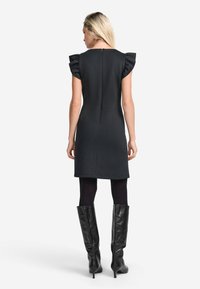Vestido negro sin mangas con mangas de volantes, forma estilizada, hasta la rodilla y con un cierre en la espalda. Combinado con botas de tacón alto negras hasta la rodilla.