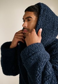 Marineblå hætteklæde i tekstureret fleece-materiale med vide ærmer og løs pasform. Stoffet virker blødt og varmt.