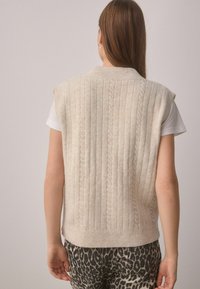 Helles beige gestricktes ärmelloses Oberteil mit vertikalem Zopfmuster und geripptem Saum, über einem weißen T-Shirt getragen. Sichtbare Leopardenmuster-Hosen.