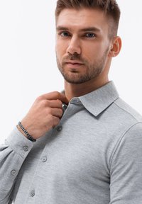 Grijze overhemd met kraag en knopen, voorzien van een zachte structuur. De stof lijkt licht van gewicht en een armband is zichtbaar om de pols.