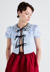 Light blue short-sleeve blouse with floral embroidery and black ribbon ties, paired with a red textured skirt featuring floral patterns.