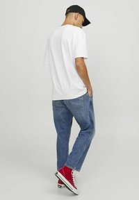 Camiseta blanca de algodón, de manga corta, con un corte casual, combinada con jeans de mezclilla azul y zapatillas altas rojas con cordones blancos. Gorra negra llevada al revés.