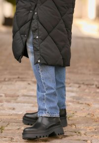 Crni kvadratičasti gležnjači s debelim potplatima i teksturiranom završnom obradom, upareni s svijetloplave ravne traperice. Tlo je od kockastog kamenja.