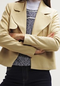 Beige imitation läder blazer med kavajkrage, strukturerade axlar och två framfickor, lager på lager över en mönstrad topp.