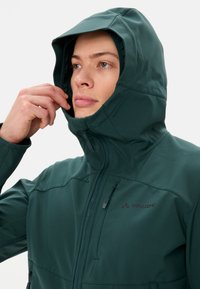 Veste softshell verte avec capuche, dotée d'une fermeture éclair à l'avant et du logo Vaude. Texture lisse et design ergonomique pour le confort.