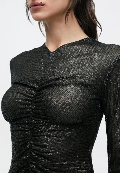 Femeie purtând un top negru cu paiete și mâneci lungi, cu detalii strânse, prezentând un prim-plan al trunchiului superior și gâtului.