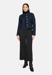 Femme portant une veste texturée bleue marine boutonnée sur un col roulé noir, un pantalon large noir et des bottes à talons noires, debout devant un fond blanc.