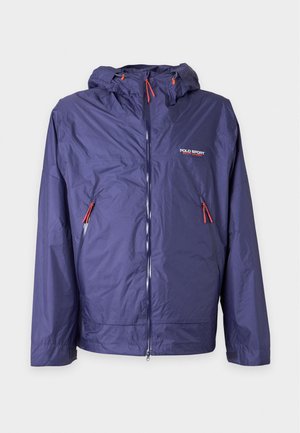 Ein junger Mann steht und trägt eine lila Kapuzen-Windjacke, dunkle weite Hosen und weiße Sneaker vor einem schlichten hellen Hintergrund.