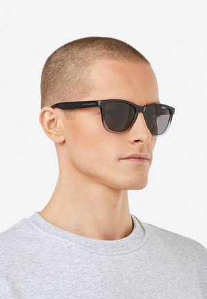 Črna plastična sončna očala s temnimi lečami, kotno zasnovo in subtilno blagovno znamko na krilu, ki jih nosi oseba v sivi jopa.