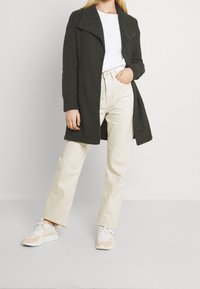 Cardigan lungo grigio scuro con colletto a scialle, indossato sopra un top bianco a coste e pantaloni beige a vita alta. Sneakers chiaro completano il look.