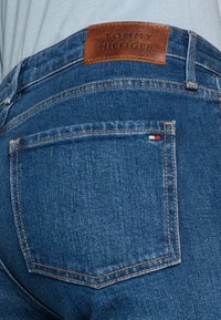 Siniset farkut, joissa on ruskea nahkaleima, jossa lukee "TOMMY HILFIGER". Farkuissa on eroavat ompeleet ja pieni brodeerattu logo taskun lähellä.